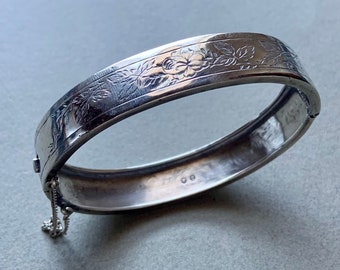 Könnte beinhalten: Ein silbernes Armreif-Armband mit floralem Design. Das Armband hat einen Scharnierverschluss und eine kleine Kette.