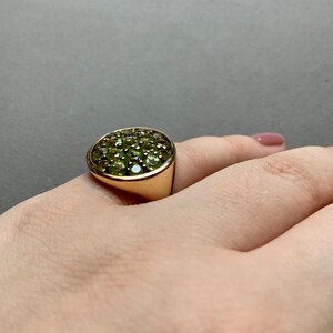 May include: A gold ring with a rounded face set with numerous small, round, green gemstones. The ring is on a person's finger, with a neutral background.