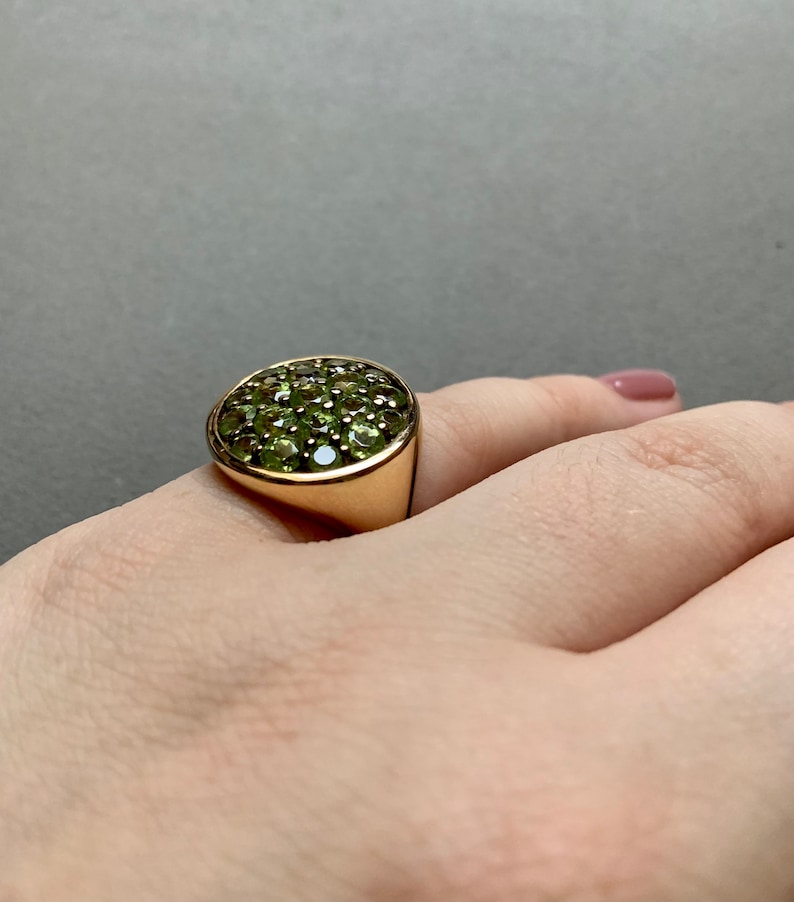 May include: A gold ring with a rounded face set with numerous small, round, green gemstones. The ring is on a person's finger, with a neutral background.