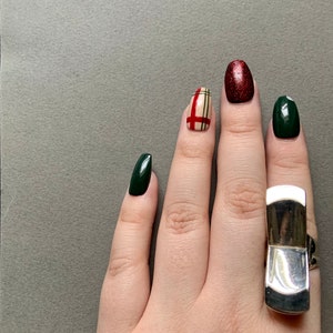 Op de afbeelding: Een hand met groene en rode nagellak, &eacute;&eacute;n nagel heeft een ruitpatroon. De hand draagt een zilveren ring.