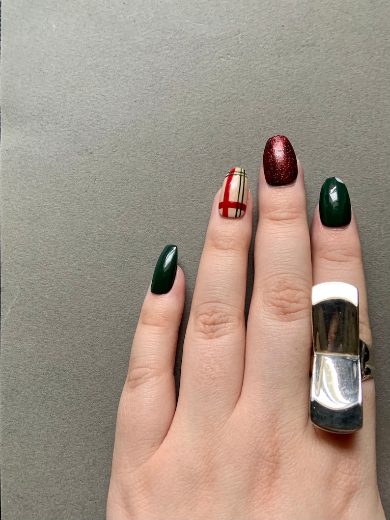 Op de afbeelding: Een hand met groene en rode nagellak, &eacute;&eacute;n nagel heeft een ruitpatroon. De hand draagt een zilveren ring.