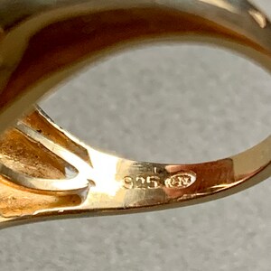 May include: Close-up of a gold-colored ring with a smooth, polished surface. The ring is stamped with the number "925" and other markings, indicating its material composition. The ring's design is simple and elegant.