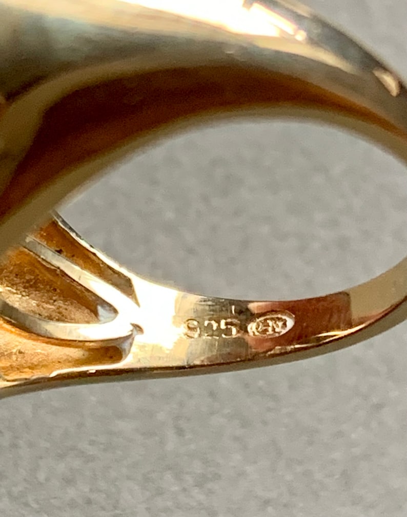 May include: Close-up of a gold-colored ring with a smooth, polished surface. The ring is stamped with the number "925" and other markings, indicating its material composition. The ring's design is simple and elegant.