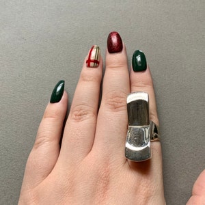 Op de afbeelding: Een zilveren ring met een uniek, abstract ontwerp. De ring wordt gedragen op de middelvinger van een hand met groene en rode nagellak.