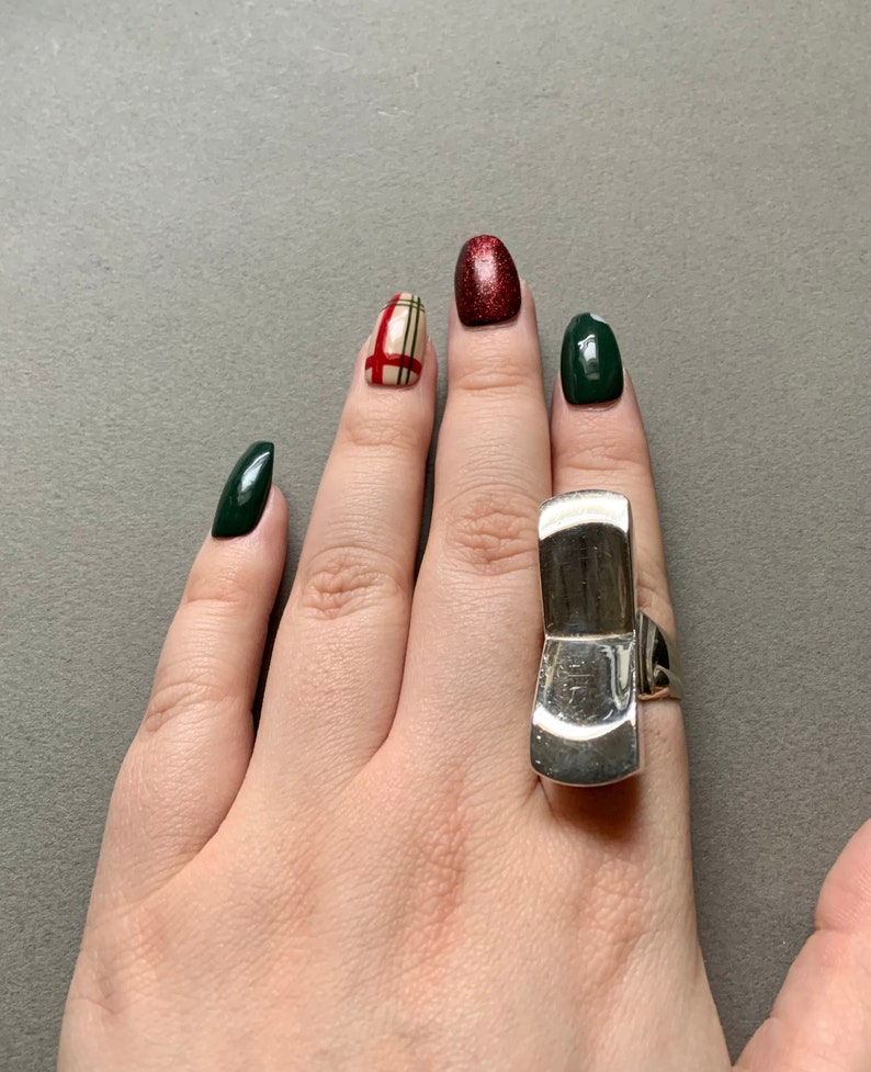 Op de afbeelding: Een zilveren ring met een uniek, abstract ontwerp. De ring wordt gedragen op de middelvinger van een hand met groene en rode nagellak.