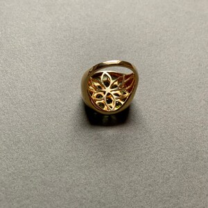 May include: A gold-colored ring with a floral filigree design. The ring has a rounded band and a large, oval-shaped face with a cut-out floral pattern. The center of the ring appears to have green gemstones.