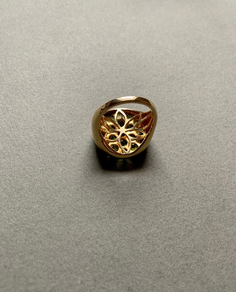 May include: A gold-colored ring with a floral filigree design. The ring has a rounded band and a large, oval-shaped face with a cut-out floral pattern. The center of the ring appears to have green gemstones.