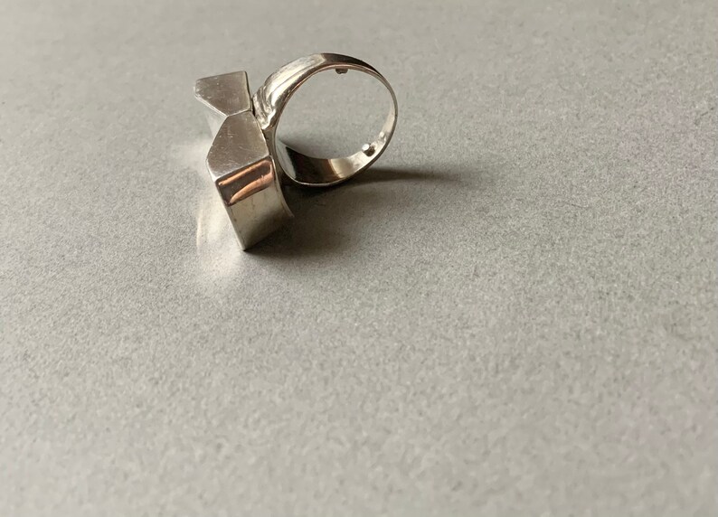 Op de afbeelding: Een zilveren ring met een geometrisch ontwerp. De ring is gemaakt van twee delen die aan elkaar zijn verbonden.