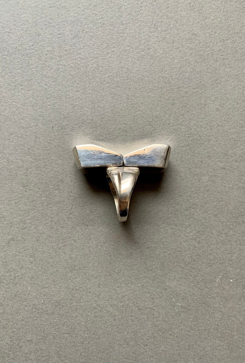 Op de afbeelding: Een zilveren ring met een uniek, geometrisch ontwerp. De ring is gemaakt van twee delen die in het midden aan elkaar zijn verbonden. De ring is een statement piece die perfect zou zijn voor een moderne bruid.