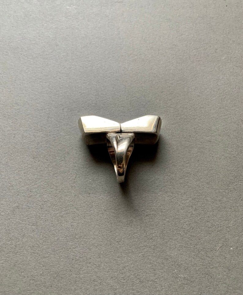 Op de afbeelding: Een zilveren ring met een uniek geometrisch ontwerp. De ring is gemaakt van twee delen die in het midden aan elkaar zijn verbonden. De ring is aan de onderkant open en heeft een gladde, gepolijste afwerking.
