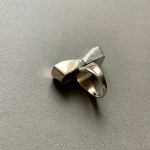 Op de afbeelding: Een zilveren ring met een uniek, geometrisch ontwerp. De ring is gemaakt van twee delen die elkaar kruisen om een boog te vormen.