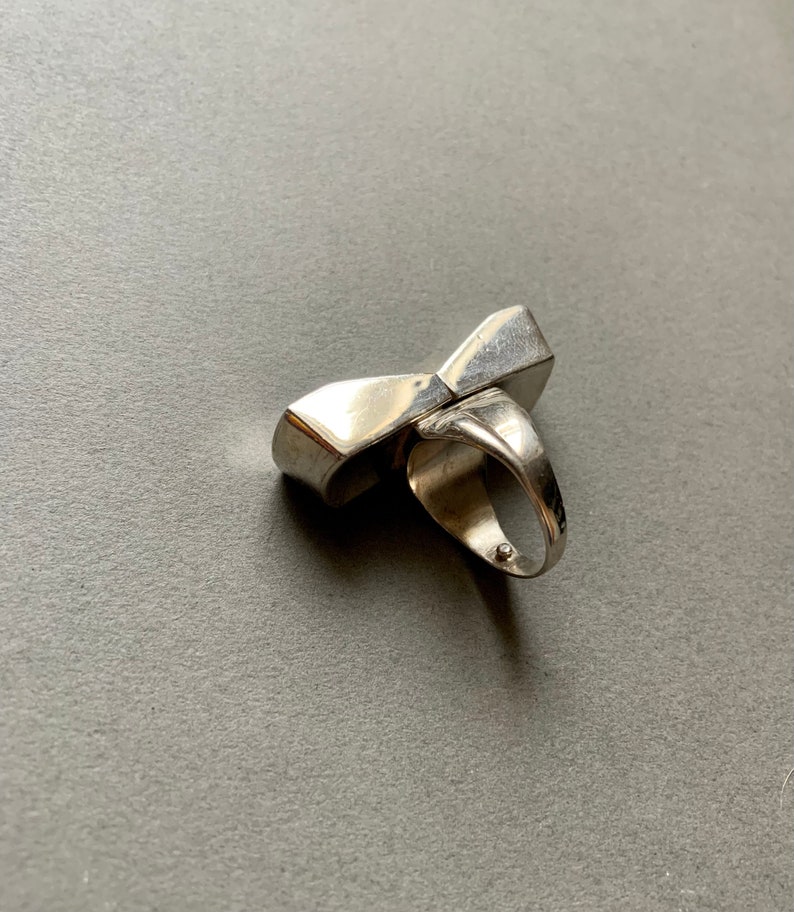 Op de afbeelding: Een zilveren ring met een uniek, geometrisch ontwerp. De ring is gemaakt van twee delen die elkaar kruisen om een boog te vormen.