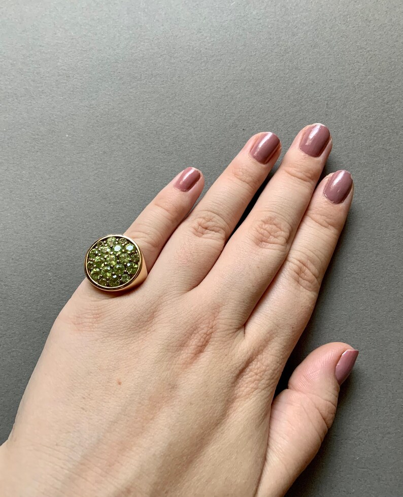 May include: A gold-toned ring with a large, round face covered in small, light green gemstones. The ring is on a hand with polished nails. The background is a solid, neutral gray.
