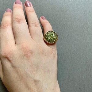 May include: A gold-toned ring with a large, circular face covered in small, light green gemstones. The ring is set on a hand with a neutral background.