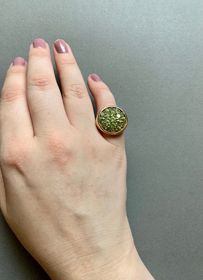 May include: A gold-toned ring with a large, circular face covered in small, light green gemstones. The ring is set on a hand with a neutral background.