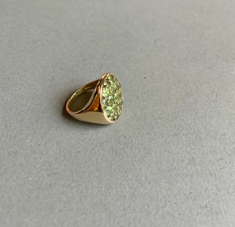 May include: A gold ring with a cluster of small, light green gemstones set in an oval shape. The ring has a smooth, polished band and a bright, reflective surface.