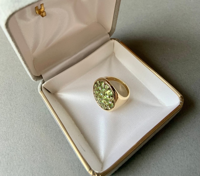 May include: A gold ring with a cluster of small, round, light green gemstones set in a circular pattern. The ring is displayed in an open, white jewelry box with a gold trim and a gold clasp.