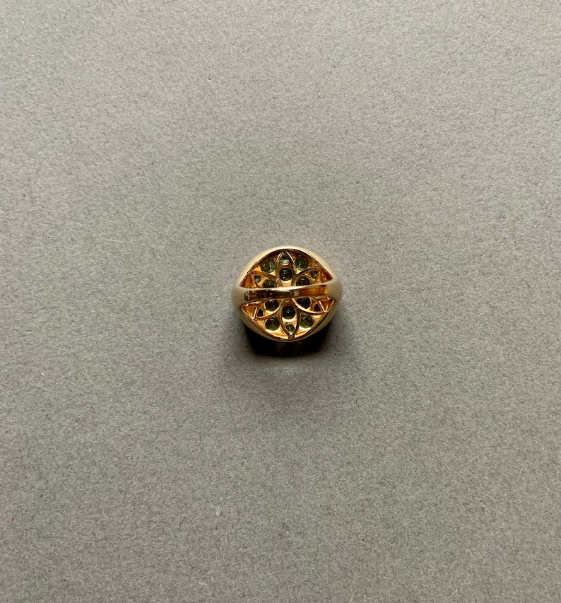 May include: A gold ring with a rounded, oval face. The face of the ring is adorned with small, dark green gemstones arranged in a floral pattern. The band of the ring is a solid, polished gold.
