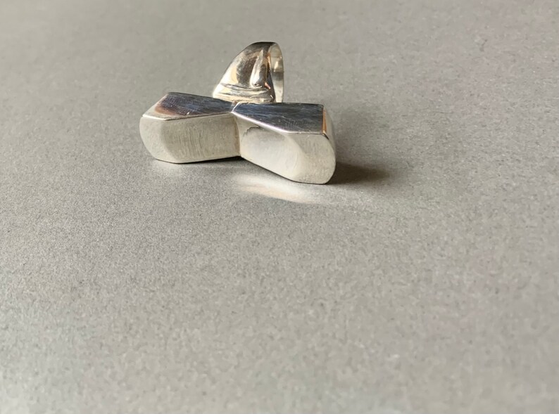 Op de afbeelding: Een zilveren ring met een geometrisch ontwerp. De ring is gemaakt van twee rechthoekige vormen die in het midden met elkaar verbonden zijn. De ring is een statement piece en zou een geweldige aanvulling zijn op elke sieradencollectie.