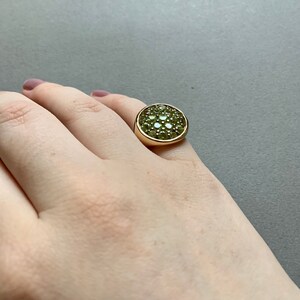 May include: A gold-toned ring featuring a large, round face covered in small, light green gemstones. The ring has a thick band and a smooth, polished finish. The gemstones are clustered closely together, creating a textured surface.