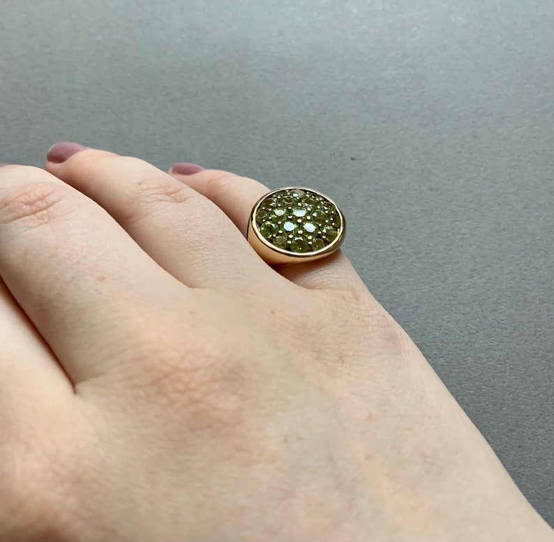 May include: A gold-toned ring featuring a large, round face covered in small, light green gemstones. The ring has a thick band and a smooth, polished finish. The gemstones are clustered closely together, creating a textured surface.