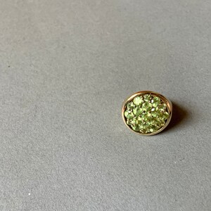 May include: A round, gold-toned ring featuring a cluster of small, light green gemstones. The ring is set against a gray background, showcasing the vibrant green stones.