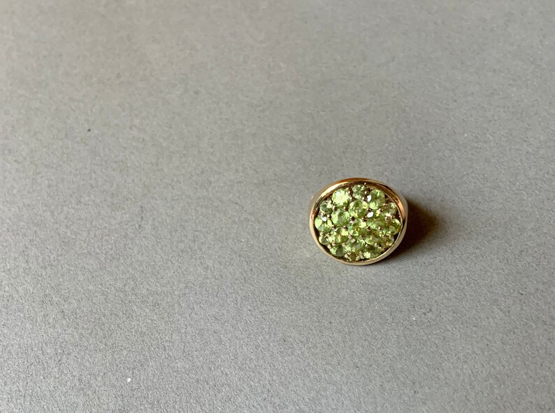 May include: A round, gold-toned ring featuring a cluster of small, light green gemstones. The ring is set against a gray background, showcasing the vibrant green stones.