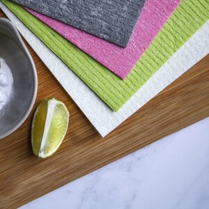 May include: A wooden cutting board with a stack of four cleaning cloths in green, pink, gray, and white. A small bowl with baking soda and two lime halves are on the board.