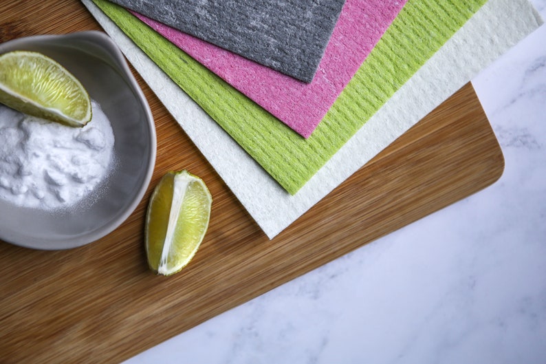 May include: A wooden cutting board with a stack of four cleaning cloths in green, pink, gray, and white. A small bowl with baking soda and two lime halves are on the board.