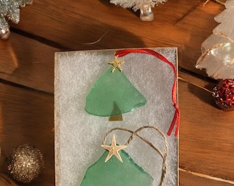 May include: Two green glass Christmas tree ornaments with starfish and gold star accents, displayed in a box with white padding. One ornament has a red ribbon, the other a twine loop. Other Christmas decorations are in the background.