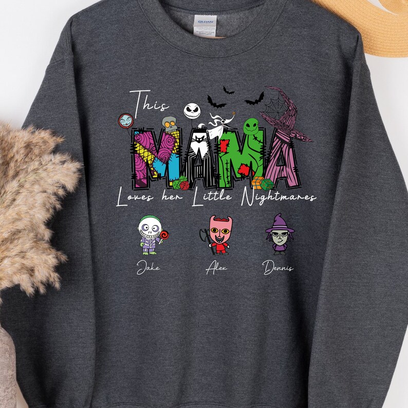 May include: A dark gray sweatshirt with a white graphic design that says "This Mama loves her little Nightmares". The graphic features a colorful "MAMA" with characters from the movie "The Nightmare Before Christmas". Below the graphic are three characters with the names "Jake", "Alex", and "Dennis" underneath.
