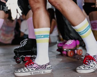 Puede incluir: Primer plano de patines y zapatillas deportivas. Las zapatillas tienen un diseño de estampado de cebra rosa y negro con cordones rojos. Se ven calcetines blancos con rayas amarillas y turquesas. También hay patines negros en la imagen.