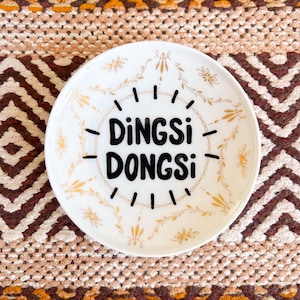 Peut inclure: Une coupelle ronde en céramique blanche ornée d'un motif en filigrane doré sur le bord, avec le texte noir "DiNGSi DONGSi" imprimé au centre, entouré d'un motif de lignes pointillées circulaires rappelant un cadran ou un soleil.