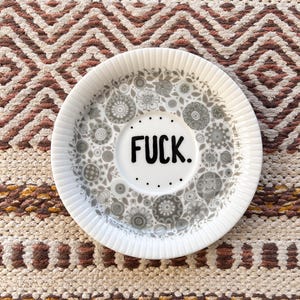 Peut inclure: Assiette en céramique blanche avec un motif floral gris et le mot "FUCK" en noir. L'assiette a un bord cannelé et est posée sur un textile à motifs.