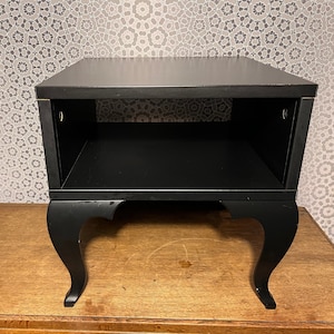 May include: A black side table featuring a flat rectangular top surface, an open storage compartment below, and four curved legs. The structure is finished in a solid black color, providing a stable base and a clear, open shelf area for storage.