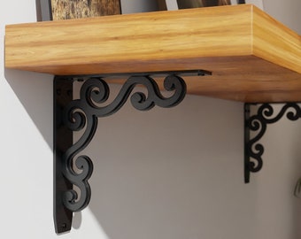 May include: Two black metal shelf brackets with a decorative scroll design are mounted to a wall. A wooden shelf with two framed pictures and a small vase with flowers is supported by the brackets.