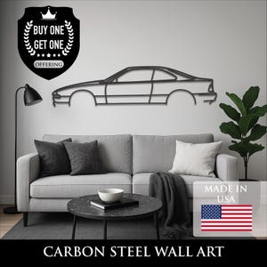 Peut inclure: Décoration murale en acier au carbone noir représentant une silhouette de voiture. L'œuvre est fixée sur un mur blanc au-dessus d'un canapé gris. L'image comprend une offre "Achetez-en un, obtenez-en un" et le texte "Made in USA".