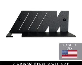 May include: Black carbon steel wall art with a geometric design. The art piece features the letters "AM" with three diagonal cutouts and two rivets. The bottom has a shelf-like extension. The text "CARBON STEEL WALL ART" is at the bottom.