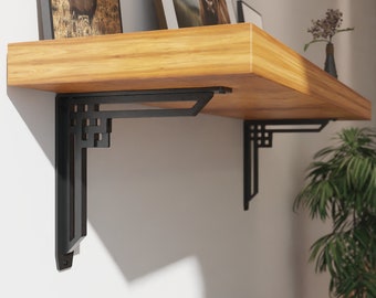 May include: Two black metal shelf brackets with a geometric design are mounted to a wall. A wooden shelf is supported by the brackets. The shelf is decorated with two framed pictures.