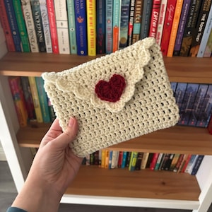 May include: A crocheted envelope-style clutch bag with a red heart applique. The bag is made with a white yarn and has a scalloped edge.