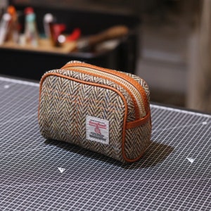 Op de afbeelding: Een kleine, rechthoekige, bruin en beige getweedde ritszak met een bruine leren afwerking. De tas heeft een gouden rits en een wit label met de tekst "Harris Tweed" in rood.
