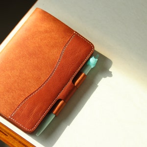 Puede incluir: Un diario encuadernado en cuero marrón con un portaplumas y una pluma azul claro. El diario tiene un detalle de costura decorativo.