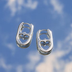 May include: A pair of silver-colored hoop earrings. Each earring has an oval shape with a heart-shaped cutout in the center. The earrings are reflective, with a bright, shiny finish. The background is a blue sky with clouds.