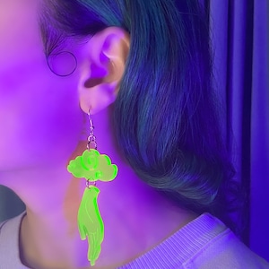 May include: Neon green acrylic earrings. Each earring features a cloud shape at the top and a hand shape dangling below. The earrings are suspended from silver-colored ear wires. The image is lit with purple and green light.
