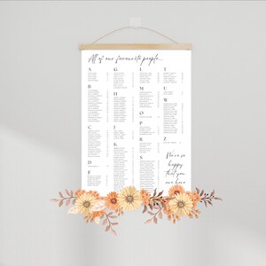 May include: A white poster with a seating chart for a wedding. The poster is hanging on a wall with a wooden frame and a rope. The poster has the text "All of our favourite people..." at the top and lists names in alphabetical order. The poster also has the text "We're so happy that you are here" at the bottom. The poster is decorated with orange and yellow flowers.