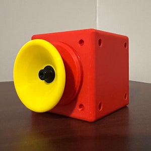 May include: A red cube with a yellow speaker attached to one side. The speaker has a black centre.