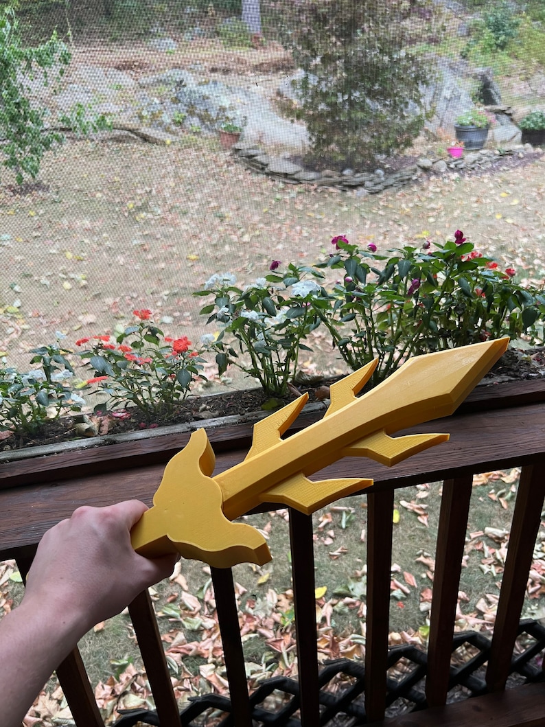 May include: A yellow plastic sword with a jagged blade and a hand holding the hilt. The sword is in front of a wooden railing with a blurry background of a garden.