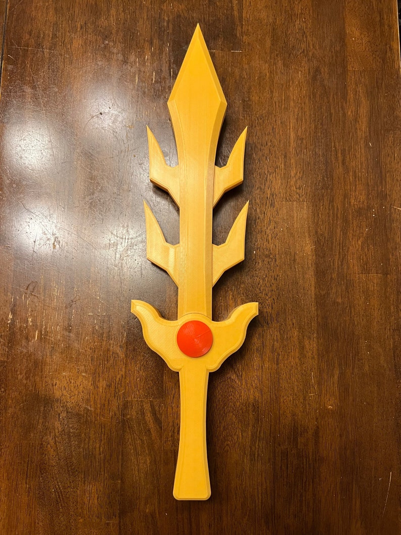 May include: A gold-colored plastic sword with a red jewel in the center of the handle. The sword has a pointed tip and a decorative design with multiple points.