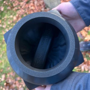 Op de afbeelding: Een donkergrijs, cilindrisch object met een getextureerde rand en een uitlopende basis. Het object heeft een holle binnenkant met een donkere, cilindrische component aan de binnenkant. Het object wordt door een persoon vastgehouden.