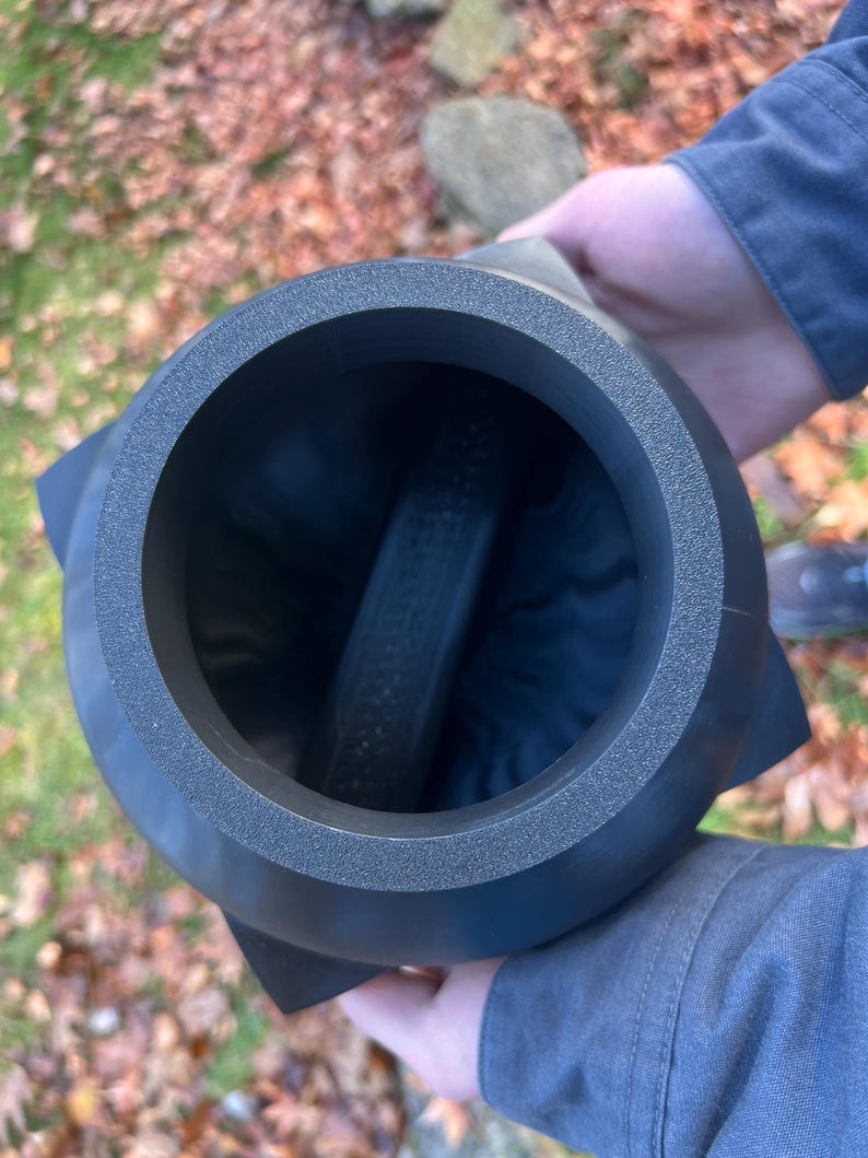 Op de afbeelding: Een donkergrijs, cilindrisch object met een getextureerde rand en een uitlopende basis. Het object heeft een holle binnenkant met een donkere, cilindrische component aan de binnenkant. Het object wordt door een persoon vastgehouden.